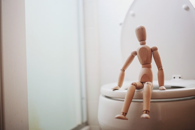 A wooden mannequin sitting on the rim of a toilet.